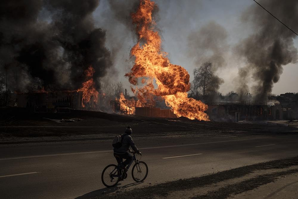 A man rides his bike past flames and smoke rising from a fire following a Russian attack in Kharkiv, Ukraine, on Friday, March 25, 2022 | AP