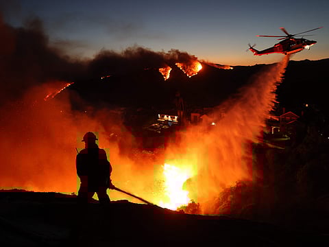 Fire personnel respond to homes destroyed while a helicopter drops water as the Palisades Fire grows in Pacific Palisades, California on January 7, 2025.  