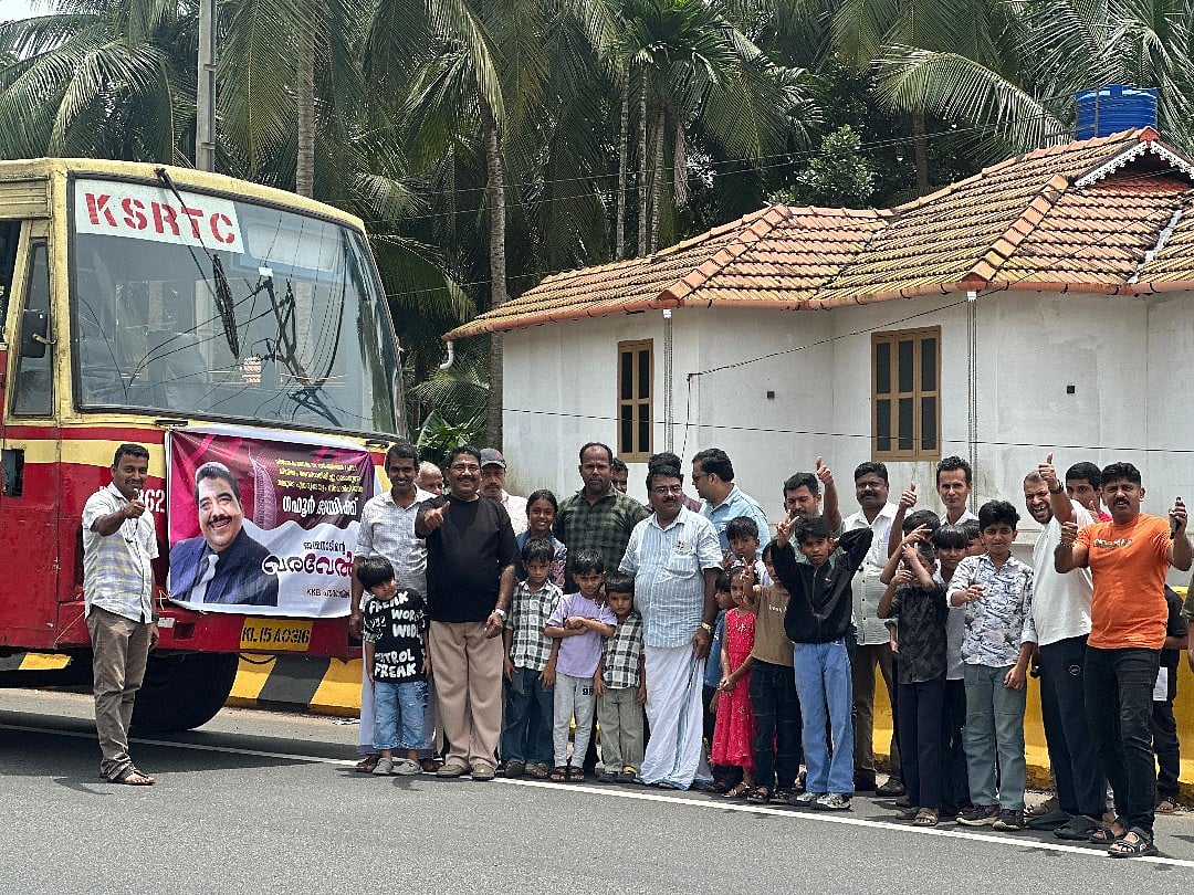 Gafoor, wearing a black t-shirt and light sandy beige pants, smiles and gives a thumbs-up alongside well-wishers by the KSRTC bus bearing a warm welcome banner.