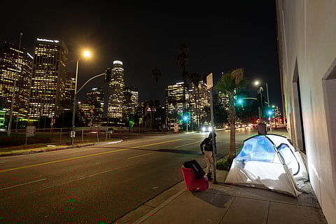 FILE - In this photo illuminated by an off-camera flash, a woman walks past a homeless person's tent with a chair in downtown Los Angeles, Feb. 16, 2023.