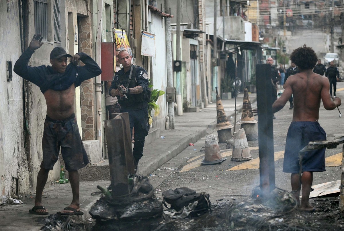A police officer holds his gun up next to residents on a barricade during the Operacao Contencao (Operation Containment) at the Vila Cruzeiro favela, in the Penha complex, in Rio de Janeiro, Brazil, on October 28, 2025.