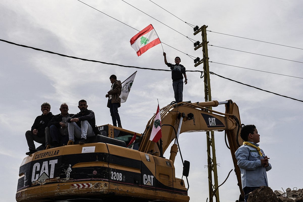 Standing on a digger used to clear the rubble of destroyed homes and businesses, a Lebanese youth holds up the Lebanese flag as residents wait for the arrival of Lebanon's prime minister during his visit to the heavily-damaged southern village of Kfar Shouba, near the border with Israel on February 8, 2026.