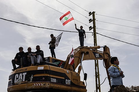 Standing on a digger used to clear the rubble of destroyed homes and businesses, a Lebanese youth holds up the Lebanese flag as residents wait for the arrival of Lebanon's prime minister during his visit to the heavily-damaged southern village of Kfar Shouba, near the border with Israel on February 8, 2026.