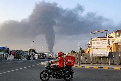 A delivery-person rides a motorcycle along a road as behind a tall smoke plume billows following an explosion in the Fujairah industrial zone on March 3, 2026.