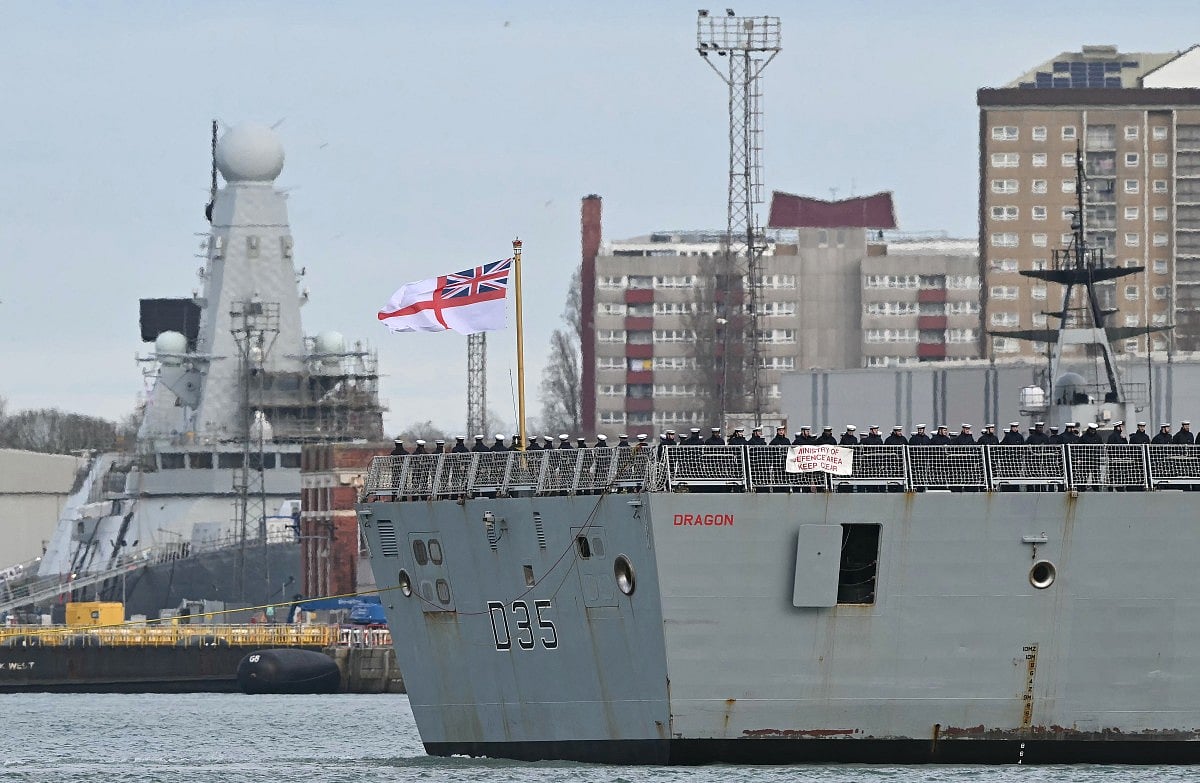 Crew members stand beneath an Ensign on the aft deck of HMS Dragon, a Royal Navy Type 45 Daring-class air-defence destroyer warship, as it is guided by tug boats from HM Naval Base Portsmouth, on the south coast of England, on March 10, 2026.