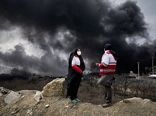 Two women from the Iranian Red Crescent Society stand as a thick plume of smoke from a U.S.-Israeli strike on an oil storage facility late Saturday rises in the sky in Tehran, Iran, Sunday, March 8, 2026. 