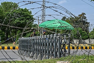Security personnel keeping watch are seen through barbed wires at a closed road leading to the Serena Hotel in the Red Zone area of Islamabad on April 23, 2026.