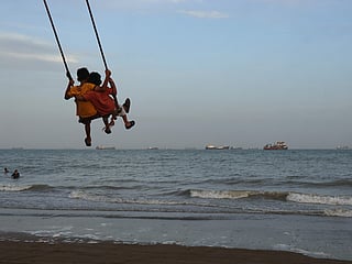 Two children swing together as a mix of bulk carriers, cargo ships, and service vessels line the horizon in the Strait of Hormuz off Bandar Abbas, Iran.