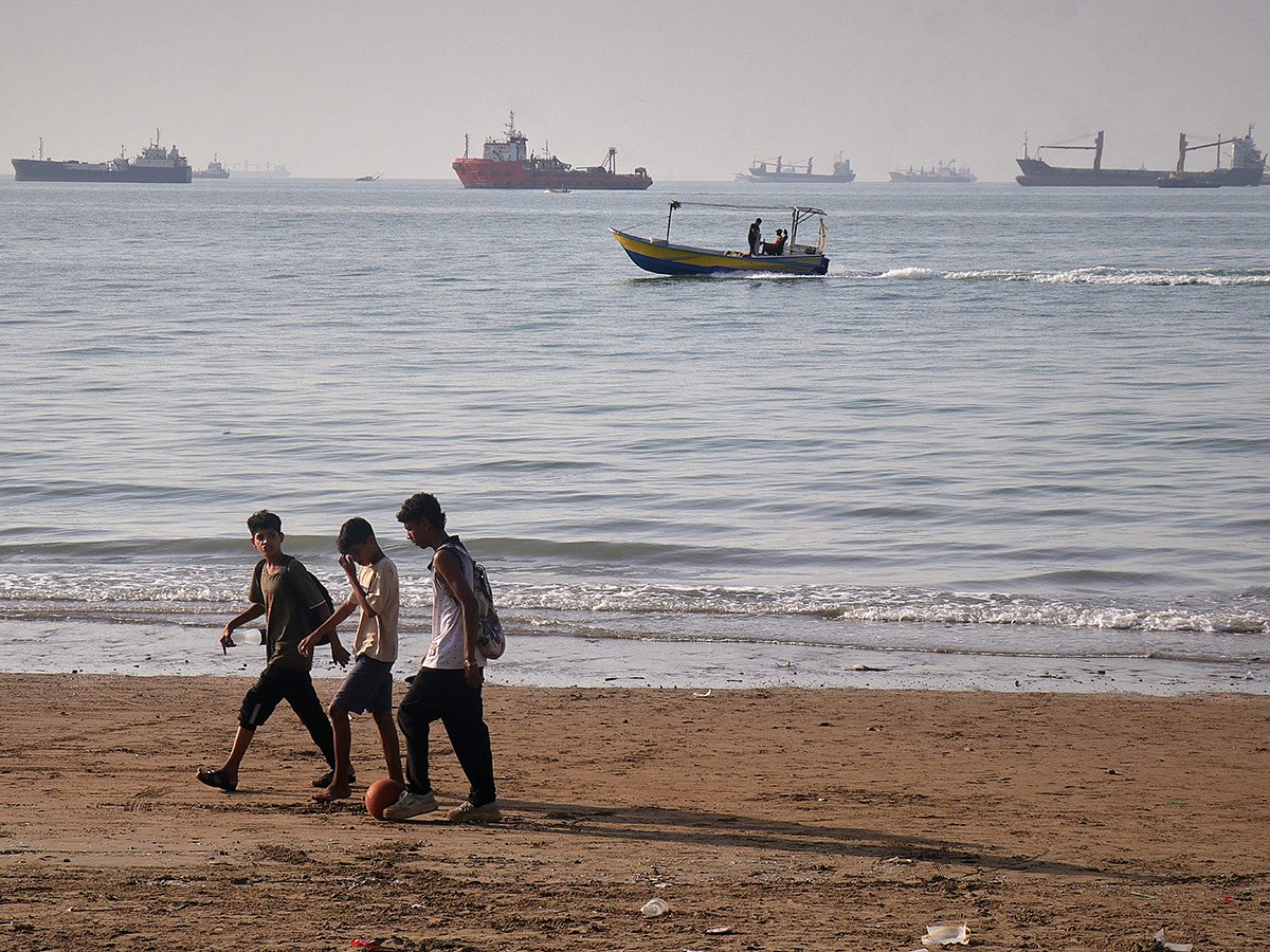 Children walk along the shore with a ball as a small motorboat passes offshore and bulk carriers, cargo ships, and service vessels line the horizon in the Strait of Hormuz off Bandar Abbas, Iran. 