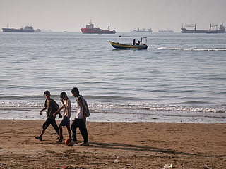 Children walk along the shore with a ball as a small motorboat passes offshore and bulk carriers, cargo ships, and service vessels line the horizon in the Strait of Hormuz off Bandar Abbas, Iran. 