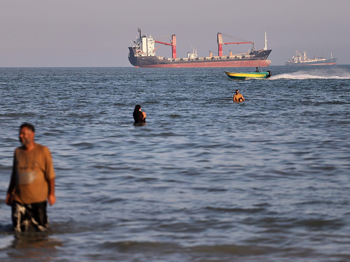 Residents swim in the waters of the Strait of Hormuz as a small motorboat passes cargo ships and other commercial vessels offshore near Bandar Abbas, in Iran. 