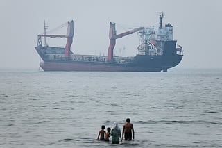 A group of people stands in shallow water as a cargo ship appears anchored in the Strait of Hormuz off Bandar Abbas, Iran. 