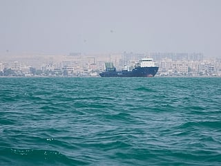 A deck cargo ship lies anchored in the Strait of Hormuz off Bandar Abbas, Iran, with the city's skyline in the background. 