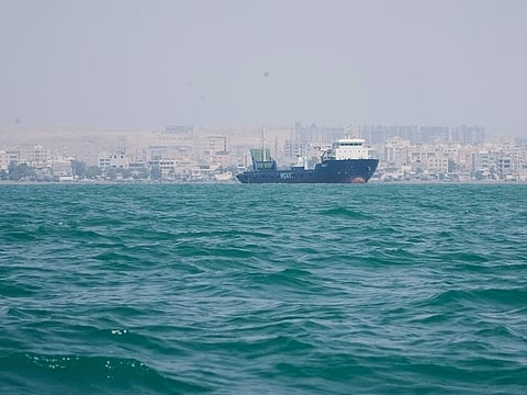 A deck cargo ship lies anchored in the Strait of Hormuz off Bandar Abbas, Iran, with the city's skyline in the background. 
