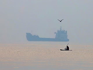 A man paddles a surfboard as a commercial vessel appears anchored in the Strait of Hormuz off Bandar Abbas, Iran. 