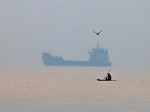 A man paddles a surfboard as a commercial vessel appears anchored in the Strait of Hormuz off Bandar Abbas, Iran. 