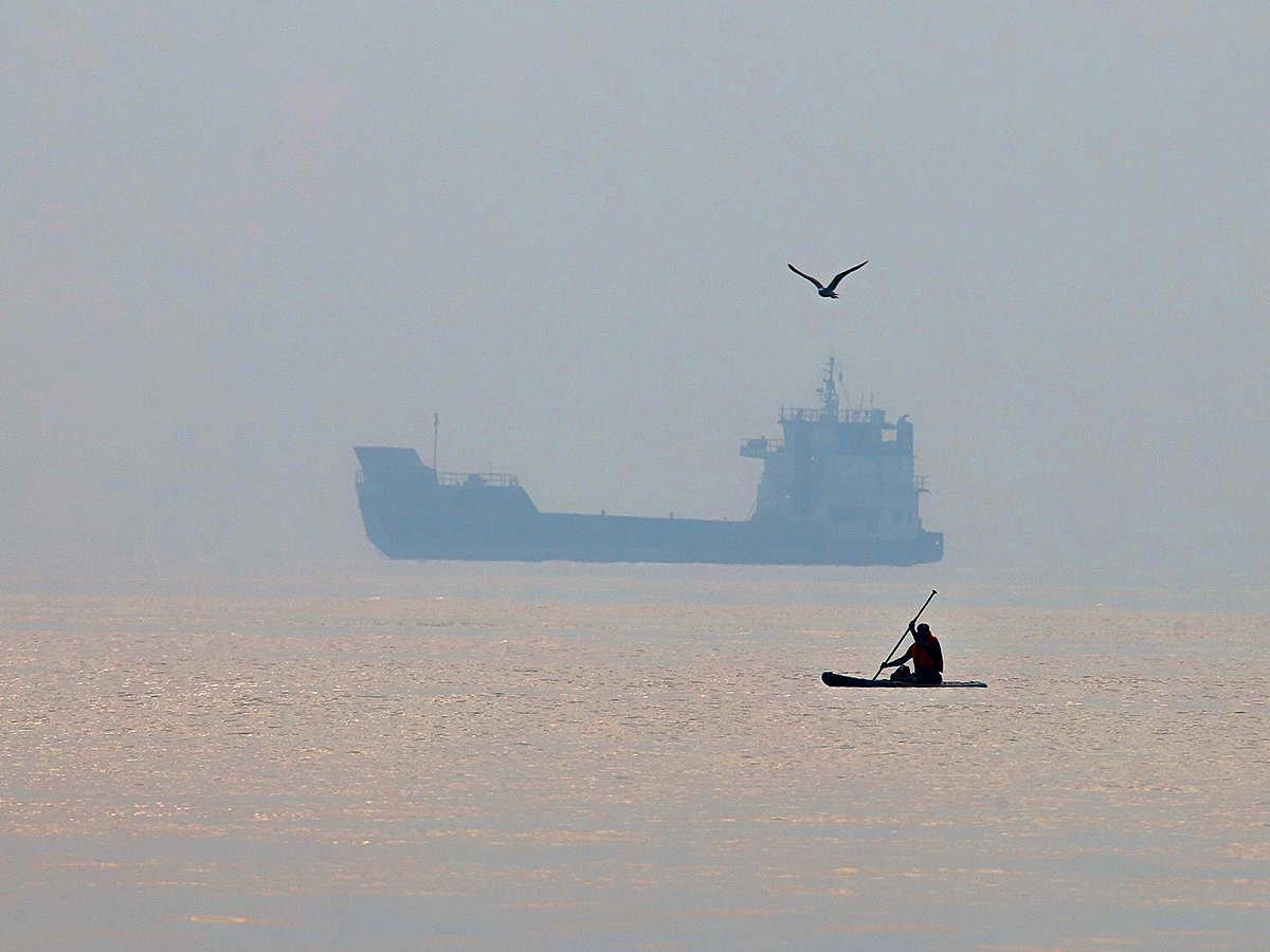 A man paddles a surfboard as a commercial vessel appears anchored in the Strait of Hormuz off Bandar Abbas, Iran. 