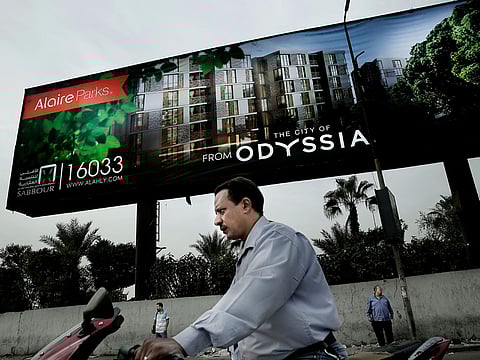 In this Oct. 23, 2018 photo, a man rides his motorbike as others wait for a bus under a residential housing compound billboard, in Cairo, Egypt.