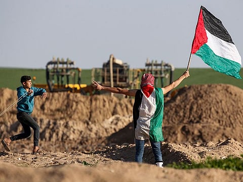 A Palestinian protester waves a Palestinian flag and flashes the victory gesture as another uses a slingshot to hurl objects during clashes following a demonstration near the border with Israel east of Gaza City