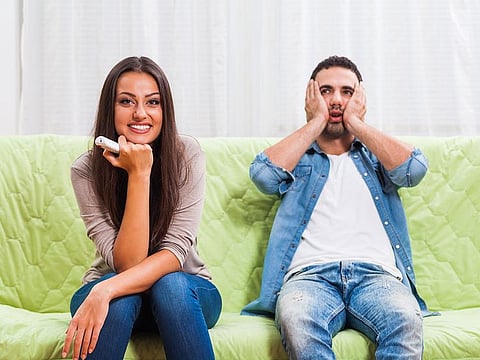 Young couple is sitting on sofa at home. Man is frustrated because he does not like what his woman is watching on TV.