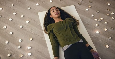 Beautiful young mixed race teenage girl with long brown hair laying on a white exercise mat in meditation while doing the yoga pose shavasana surrounded by candles in a dimly lit yoga studio.