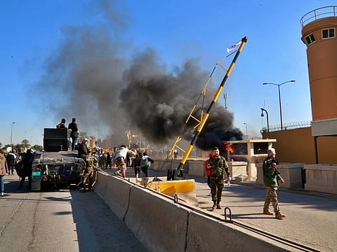 Security forces stand guard while protesters burn property in front of the US embassy compound, in Baghdad Tuesday.