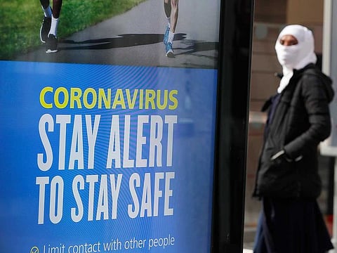 A woman with her face covered waits at a bus stop in London, Tuesday, May 12, 2020, as the country continues in lockdown. Britain's Prime Minister Boris Johnson announced Sunday that people could return to work if they could not work from home.