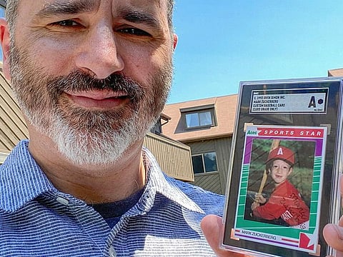 Allie Tarantino holding a baseball card featuring a very young Mark Zuckerberg grinning in a red jersey and gripping a bat. For 30 years Tarantino kept the baseball card filed it away in his basement, not knowing Zuckerberg would someday become a household name. 