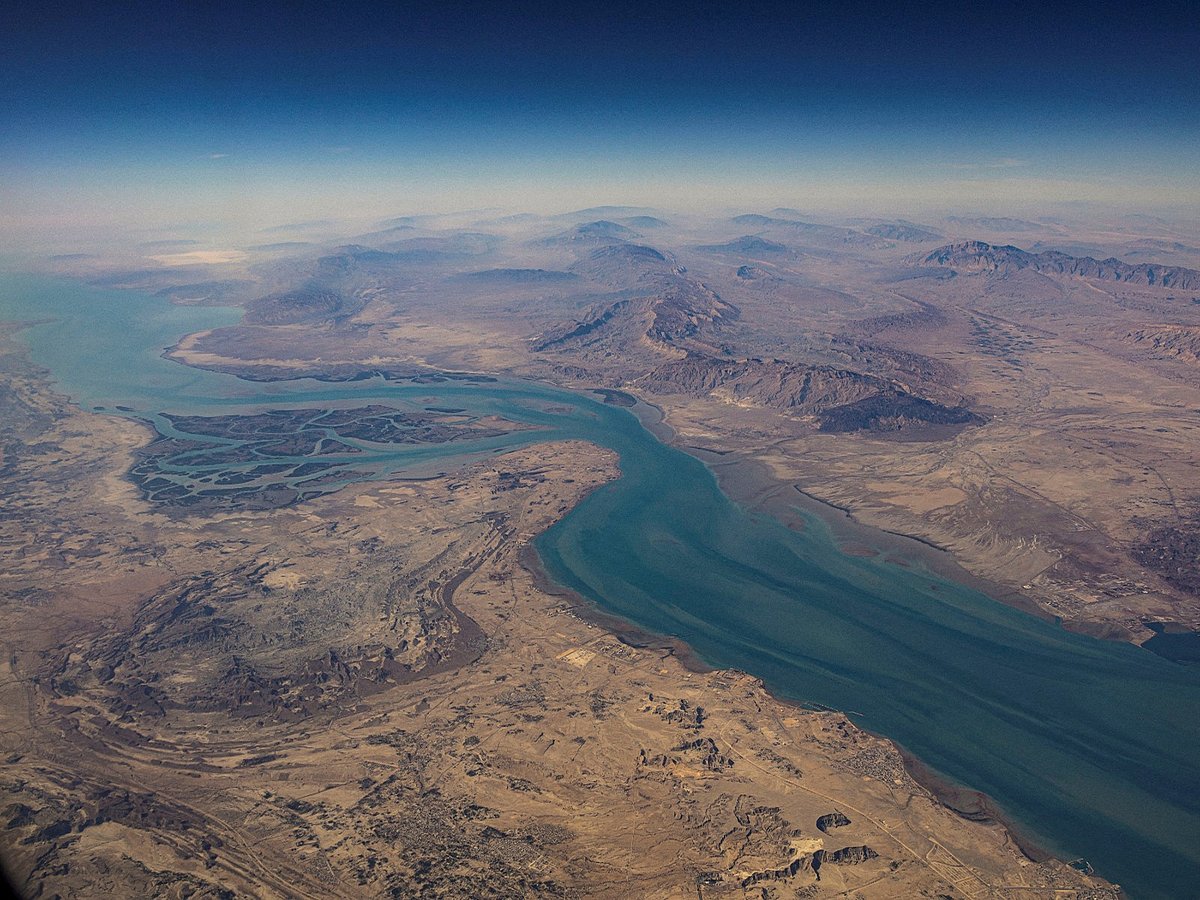 A large cargo ship carrying colorful shipping containers navigates through a narrow blue sea channel.