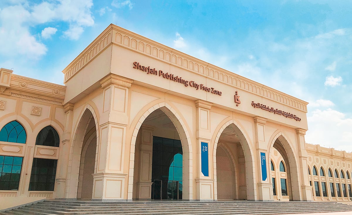 A modern office building facade with glass windows reflects the bright blue sky above the Sharjah city skyline.