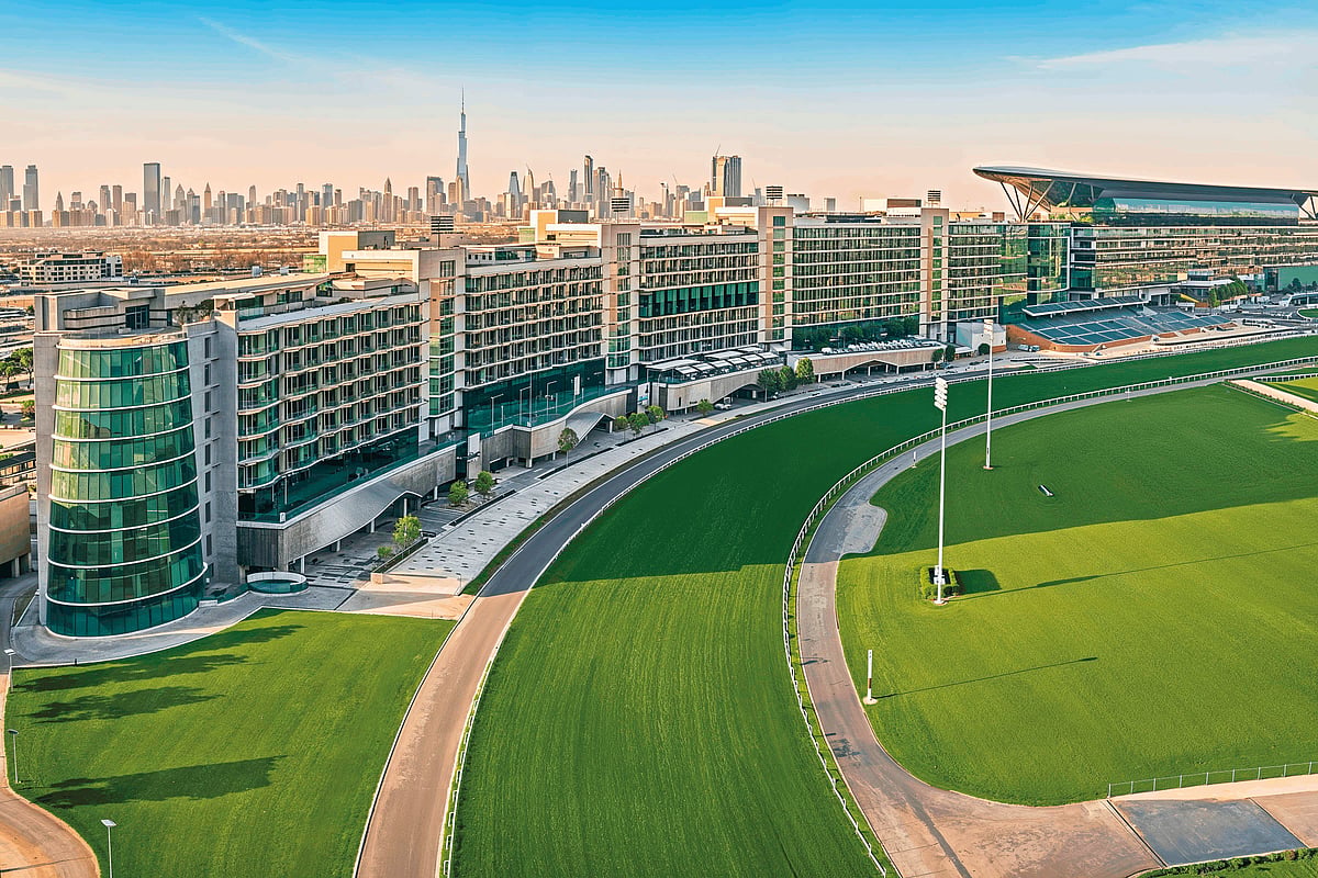 A modern glass skyscraper rises above a manicured landscape in Dubai's business district under a clear blue sky.