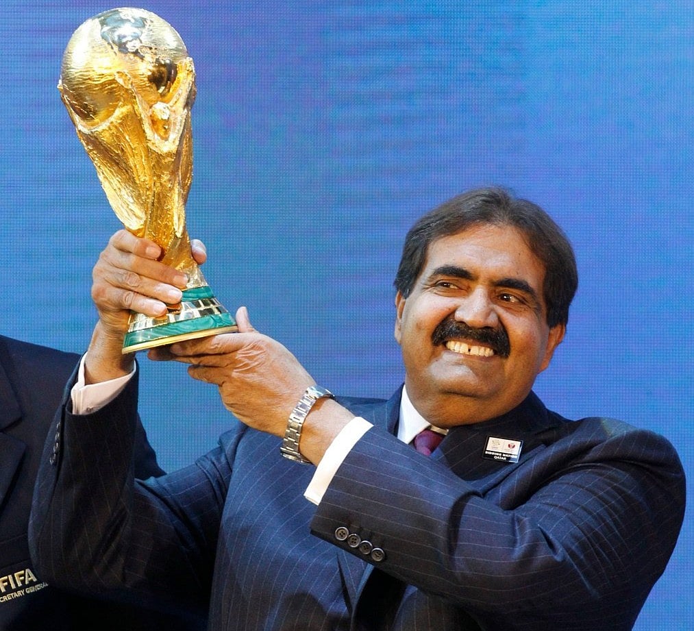 Sheikh Hamad bin Khalifa Al Thani stands formally in traditional white Qatari attire against a neutral backdrop.