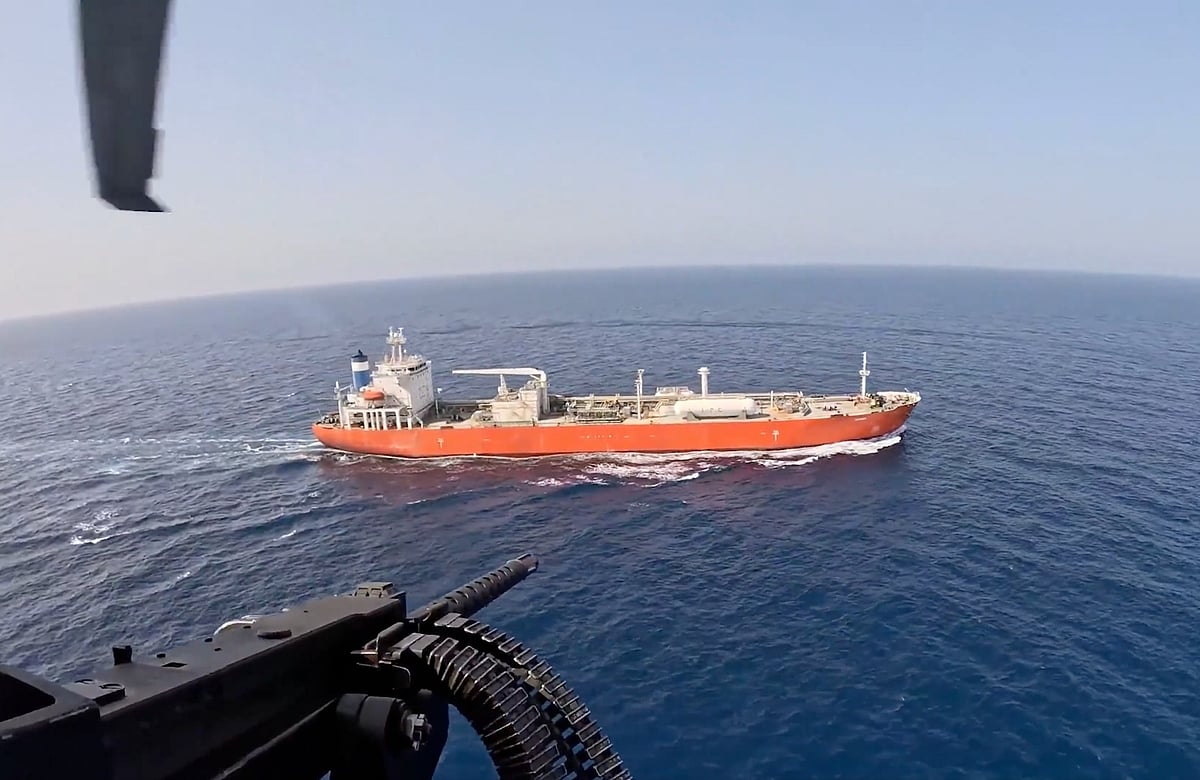 A cargo ship sails through the narrow Strait of Hormuz as thick black smoke rises from the distant horizon.