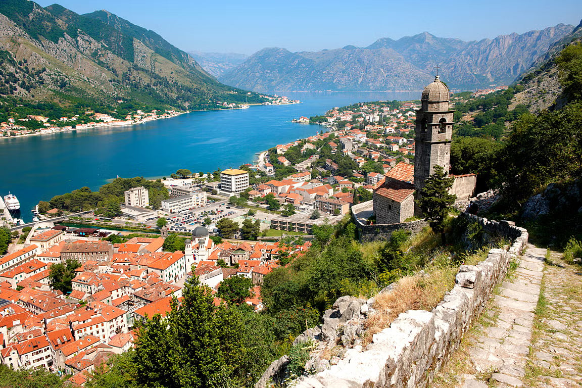 A historic stone village with orange-tiled roofs sits on a small island surrounded by calm blue Adriatic waters.
