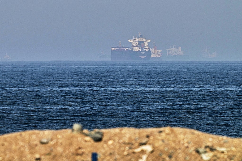 A large oil tanker sails through the calm blue waters of the Strait of Hormuz under a clear, bright sky.
