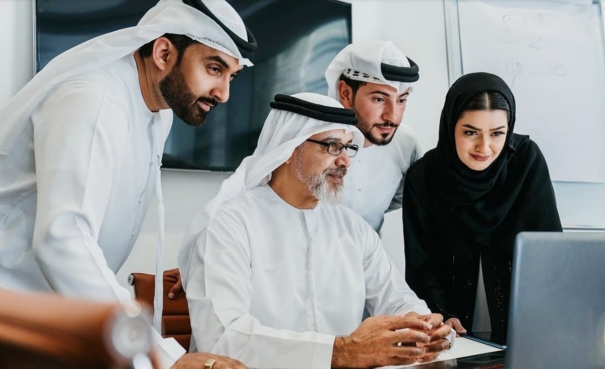 A group of professional Emirati office workers engages in a business discussion within a modern corporate workspace.
