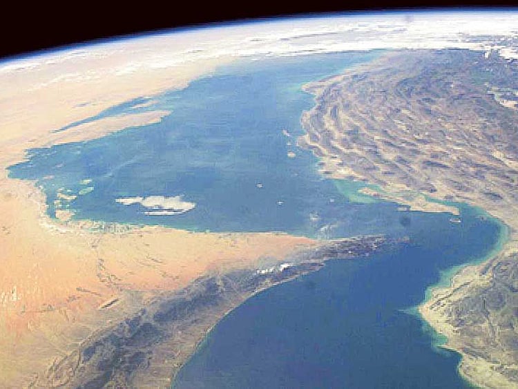 Cargo ships navigate the blue waters of the Strait of Hormuz beneath rugged, desert mountains on the coastline.
