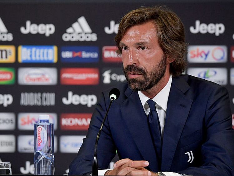 Andrea Pirlo stands on a football pitch wearing a navy tracksuit while coaching players during a daytime training session.