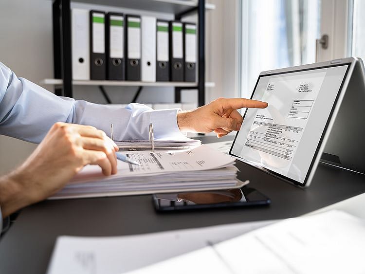 A small business owner uses a laptop and calculator to organize financial documents and tax forms at a wooden desk.