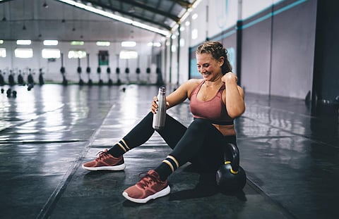 Cheerful fit girl dressed in tracksuit holding water bottle in hand and smiling during training day in gym, happy Caucaisan woman with protein cocktail resting in sportive studio - healthy lifestyle