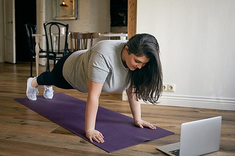 Energetic self determined overweight young woman in sneakers standing in plank with feet and hands on mat, reteating exercises after professional fitness instructor, watching tutorial online on laptop
