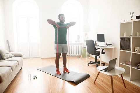 Healthy life. Full length shot of male fitness instructor showing exercises with resistance band, streaming, broadcasting video lesson on training at home using laptop. Sport, online gym concept