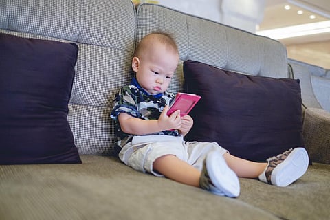 Cute little Asian 18 months / 1 year old baby boy child sitting on sofa watching a video from smart phone. Kids playing with smartphone. leisure & children & technology & internet addiction concept