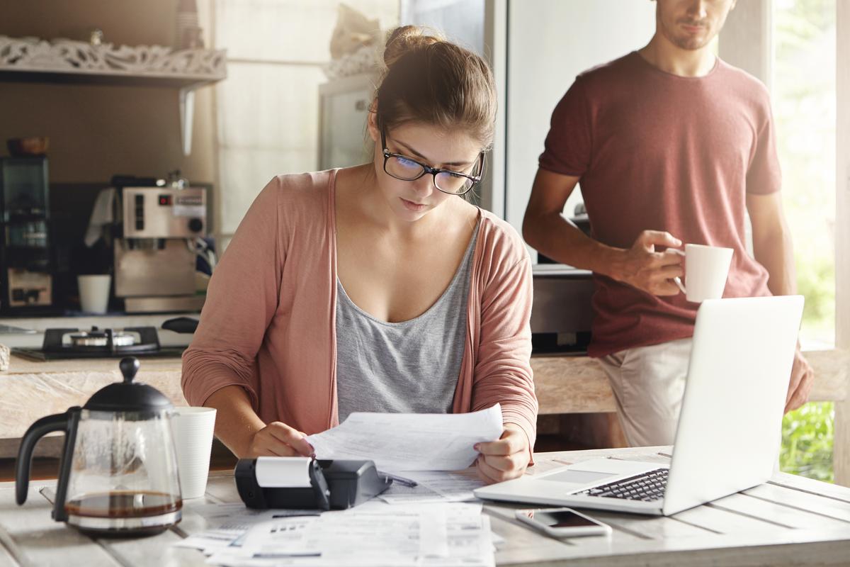 Mujer joven haciendo cuentas junto con su esposo en casa, planeando una nueva compra. Mujer seria con gafas sosteniendo un pedazo de papel y haciendo los cálculos necesarios