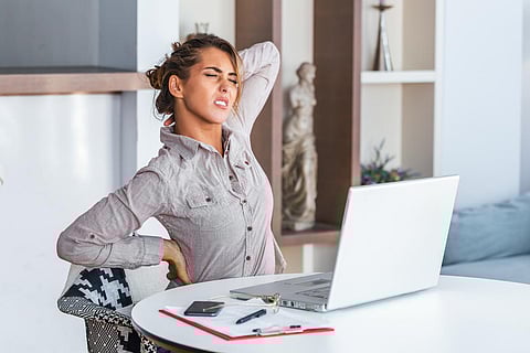Portrait of young stressed woman sitting at home office desk in front of laptop, touching aching neck with pained expression, suffering from neck pain after working on pc