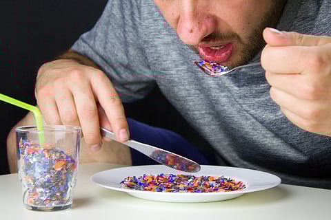 The man eats pieces of crushed recycled plastic from white plate.Food and drink contains plastics. A glass full of recycled plastic and a plastic pieces on white plate.