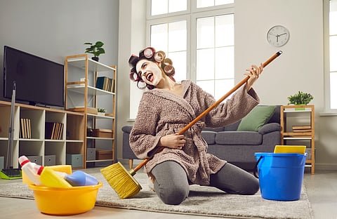 Cheerful young woman with musical talent having fun while cleaning her house. Funny crazy housewife in hair curlers and face mask tidying up home, singing loud songs and playing on pretend mop guitar
