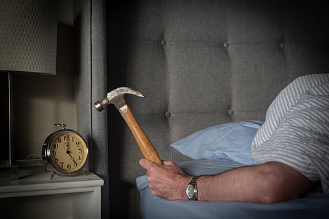 Person sleeping in bed about to strike their ringing morning alarm clock with a hammer.