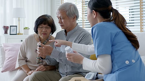 Young caregiver in scrubs uniform showing medicine bottle to elderly asian couple man and woman in home visit care nursing service. Asian senior with assisted living medication monitoring concept.