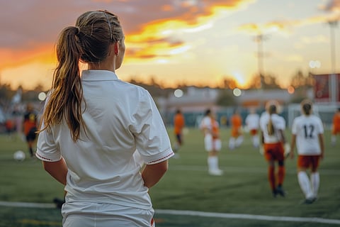 Female soccer coach in white shirt watches team play.